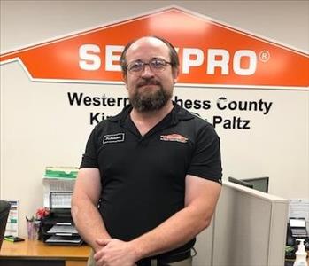 A male employee standing in front of the SERVPRO Orange House logo, wearing a SERVPRO shirt