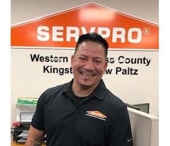 A smiling male employee standing in front of the SERVPRO Orange House logo, wearing a blackSERVPRO shirt