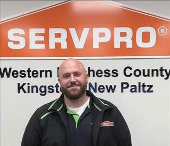 A photo of a smiling employee in a black jacket standing in front of the SERVPRO orange house logo