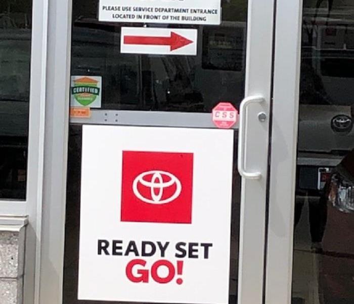 A photo of glass doors of a car dealership displaying the Certified: SERVPRO Cleaned badge
