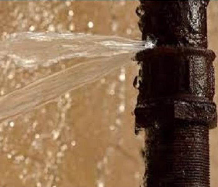 A close up of a vertical-standing, rusty pipe spraying water from a pin-hole break in it.
