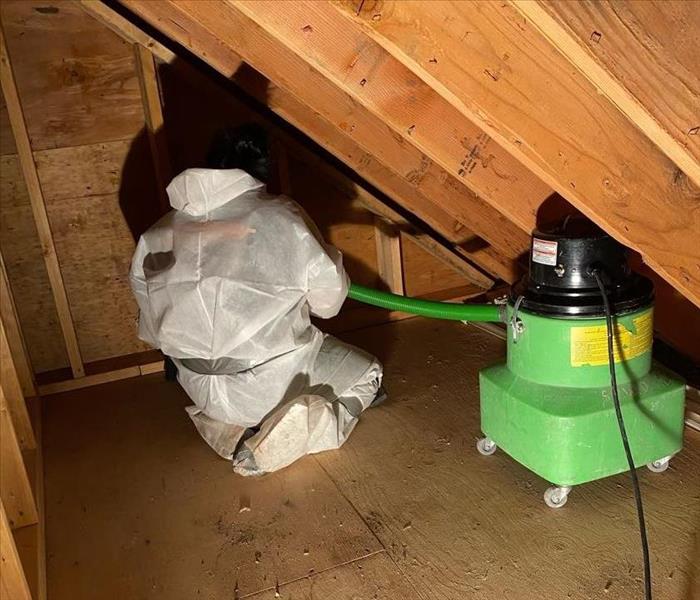 A SERVPRO employee in white PPE using a hepa-vacuum to clean remaining debris in an attic.